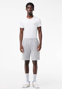 Jeune homme debout portant un t-shirt blanc, un short gris, des chaussettes blanches et des baskets blanches sur un fond blanc uni.
