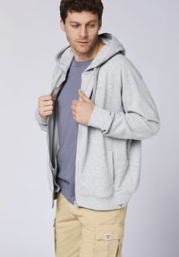 Gray zip-up hoodie made of soft fabric, featuring a front zipper, two side pockets, and ribbed cuffs, worn over a blue shirt and beige cargo shorts.