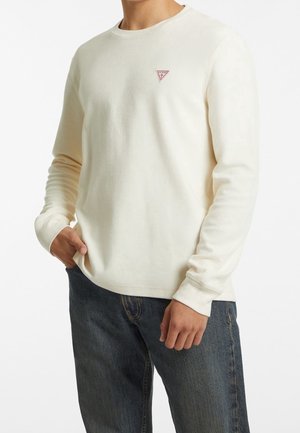 Homme portant une chemise crème à manches longues avec un petit logo triangulaire rouge sur la poitrine et un jean bleu foncé, main dans la poche.