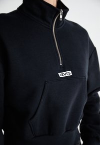 Felpa nera con zip e colletto alto, tasca frontale e patch con logo "LEVI'S" bianco. Presenta una texture morbida e un elastico in vita aderente.