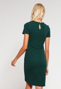 Kobieta o blond włosach ubrana w dopasowaną, krótko rękawową, ciemnozieloną sukienkę do kolan, pokazana od tyłu z detalem w kształcie klucza przy szyi.