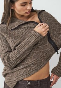 Bruine gebreide pullover met rits, voorzien van geribde mouwen en textuurpatronen. Relaxte pasvorm, waardoor een hint van huid bij de taille zichtbaar is.