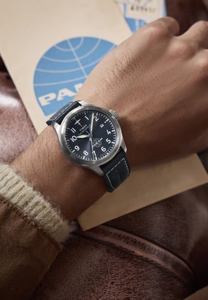 Zegarek Timex z srebrną stalową kopertą, czarną tarczą, białymi cyframi i ciemnym skórzanym paskiem. W tle widać brązową skórzaną powierzchnię.