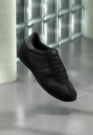 Sapato atlético preto com parte superior em couro liso e malha, fecho com atacadores e marcações discretas na lateral e na língua. Sola de borracha.