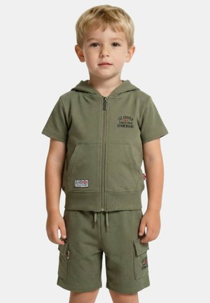 Jeune garçon portant un sweat à capuche zippé à manches courtes vert olive et un short cargo assorti avec des logos de marque, debout devant un fond uni.