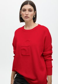 Roter übergroßer Pullover mit prägenden Logo-Details auf der Vorderseite, gerippten Ärmeln und rundem Ausschnitt. Hergestellt aus weichem Stoff mit glatter Textur.
