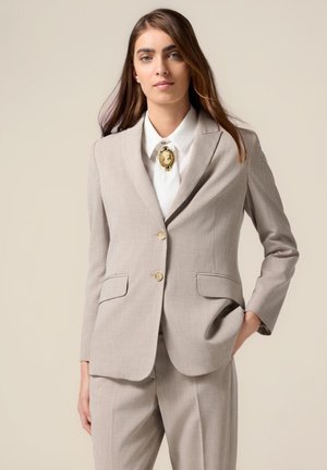 Giacca da completo beige chiaro con due tasche frontali, bottoni dorati e rever a pistagna, abbinata a una camicia bianca e a una spilla decorativa.