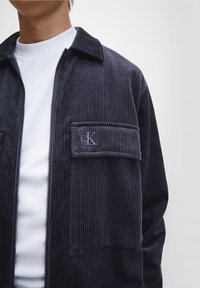 Marineblaue Cordjacke mit Reißverschluss vorne, knöpfbarem Kragen und aufgesetzter Tasche mit gesticktem CK-Logo. Trägt man über einem weißen, strukturierten Hemd.