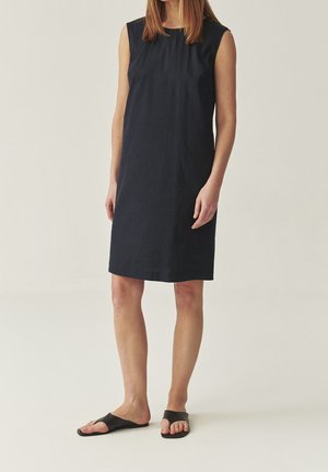 Femme portant une robe noire sans manches, arrivant au genou, et des sandales noires à enfiler, se tenant devant un fond clair et uni.