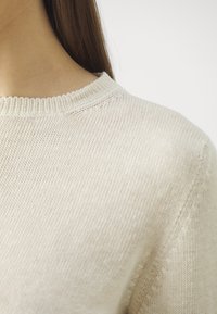 Pull beige clair à encolure ronde, fabriqué en tissu maille doux, présentant une texture subtile et une bordure côtelée délicate au niveau du col.