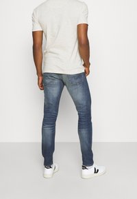 Modré džíny ze strečového denimu s úzkým střihem, zadními kapsami a mírným vyblednutím. Kombinovány se světle šedým tričkem a bílými teniskami.