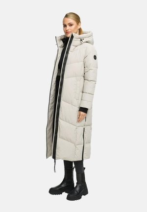 Woman wearing long beige padded coat with hood, black pants, and black chunky boots, standing against a white background.