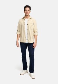 Camisa de pana de color beige claro con botones negros, llevada sobre una camiseta blanca. Jeans de color azul oscuro y zapatillas blancas completan el atuendo.