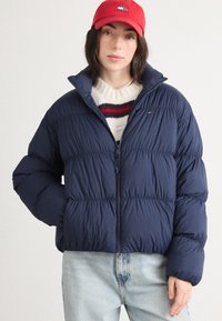 Granatowa kurtka puchowa z wysokim kołnierzem, zamkiem błyskawicznym i segmentowanymi przeszyciami, w połączeniu z czerwoną czapką i swetrem w kremowe paski.