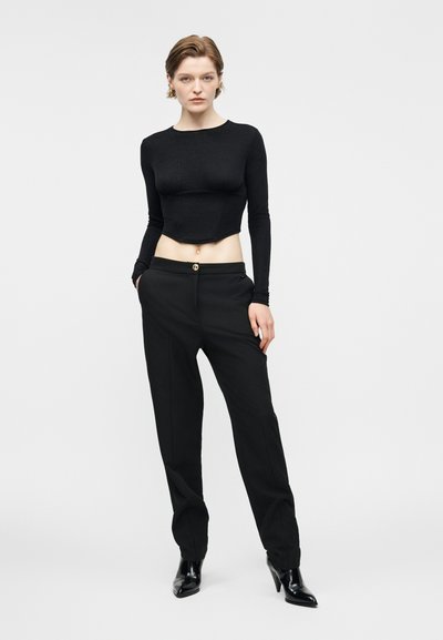 Femme portant un crop top noir moulant à manches longues, un pantalon noir taille haute et des bottines noires, se tenant avec une main dans la poche.