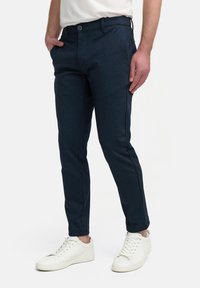 Marineblauwe slim-fit broek van een gestructureerde stof, met zakken aan de voorkant en een knoopsluiting, gecombineerd met witte sneakers.