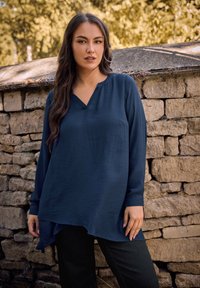Blusa lunga a maniche con scollo a V di colore blu navy, realizzata in tessuto leggero. Presenta una vestibilità ampia con orlo curva e una texture liscia.