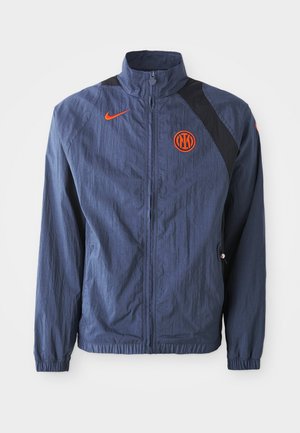 Tumš zilas vējjaka ar melnu plecu paneli, rāvējslēdzēju priekšpusē, apkakli, elastīgiem aprocēm un oranžu Nike logo un emblemu akcentiem.