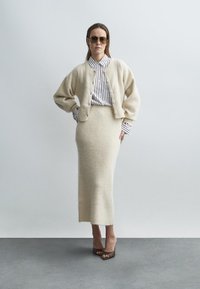 Completo cardigan e gonna color crema con finitura morbida e testurizzata. Camicia a righe con bottoni sotto. Modella indossa sandali marroni con tacco alto e occhiali da sole.