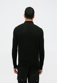 Juodas megztinis su ilgomis rankovėmis, su raštuotu apykakle ir tiesiu siūlėmis. Paviršius atrodo lygus ir minkštas.