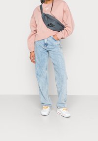 Ljusrosa sweatshirt, grå nylonmidjeväska med svart logotyp, ljusblå high-waisted jeans och vita sneakers med orange detaljer.