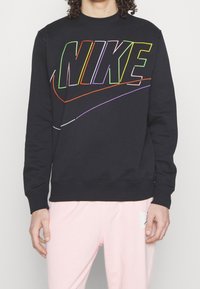 Person som bär en svart sweatshirt med färgglad kontur av "NIKE"-texten och swoosh-logon, i kombination med ljusrosa byxor.
