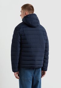 Marineblaue Steppjacke mit Kapuze, ausgestattet mit horizontalen Steppnähten und einer lockeren Passform. Kombiniert mit blauen Jeans.