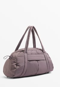 Sac de sport rembourré en mauve doux, doté de poignées doubles, d'une poche zippée et d'une forme arrondie avec un tissu texturé.