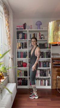 Grå halterneck jumpsuit med dyb V-udskæring, parret med multicolor sneakers. Baggrunden viser bogreoler, planter og dekorative genstande.