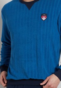 Blue textured sweatshirt featuring vertical ribbing, a navy round neckline, and a patch with red and white detailing on the chest.