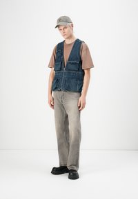 Vest din denim cu buzunare frontale peste un tricou maro, asortat cu blugi gri drepti și pantofi masivi negri. Fundal deschis.