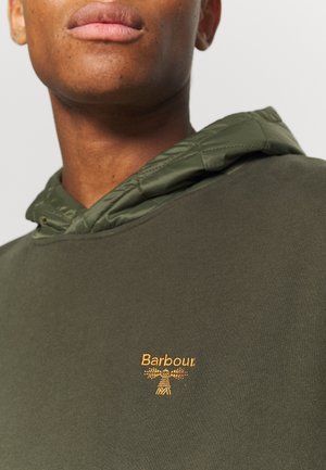 Zbliżenie na osobę noszącą oliwkowo-zieloną bluzę z kapturem Barbour z pikowanym detalem kaptura i haftowanym żółtym logo Barbour na piersi.