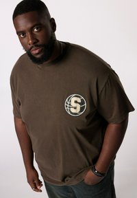 Brun bomulds T-shirt med korte ærmer, prydet med et tekstureret rundt logo med et stiliseret "S" i midten og en rund hals.