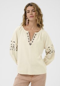 Blouse de couleur crème avec un décolleté en V, ornée de broderies florales en rose et bleu, et de longues manches ballons avec des motifs tribaux près des poignets.