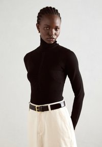 Zwarte coltrui met ribbelstructuur, aangesloten silhouet en geplooide details aan de voorkant, gecombineerd met hoogtailleren beige broeken en een zwarte riem.