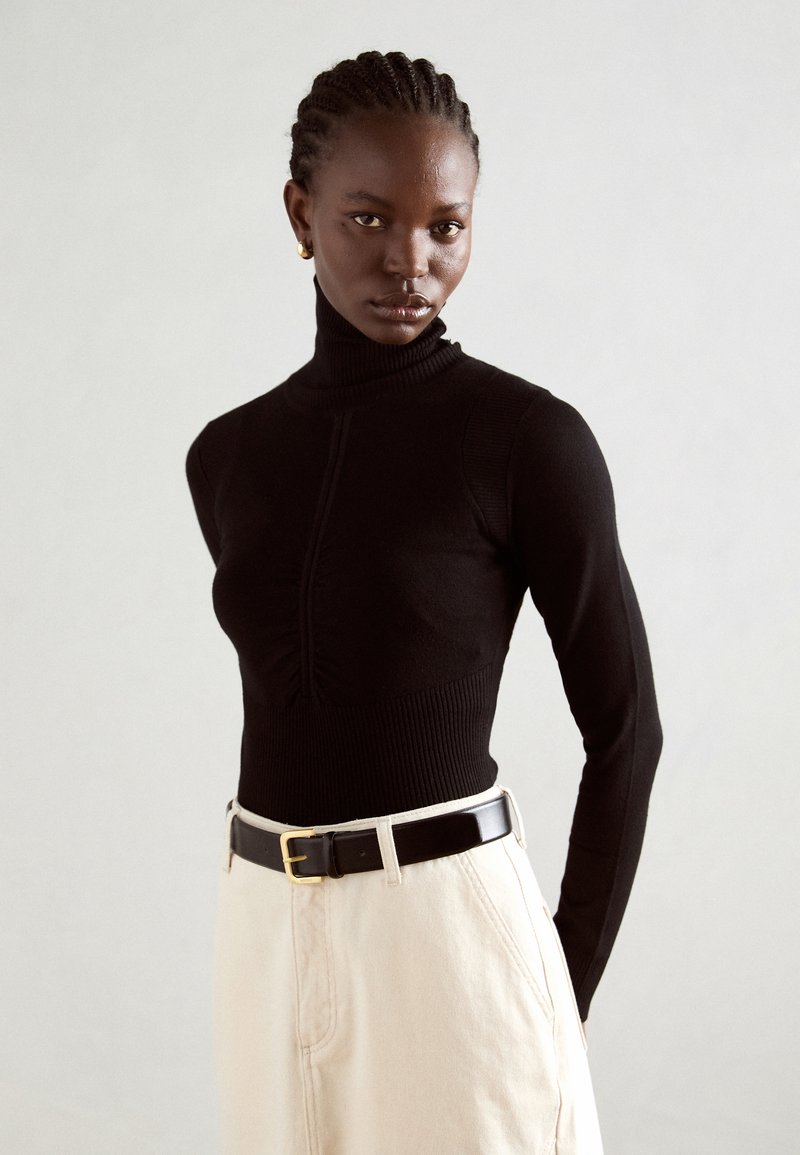 Maglione a collo alto nero con texture a costine, silhouette aderente e dettagli arricciati sul davanti, abbinato a pantaloni beige a vita alta e una cintura nera.