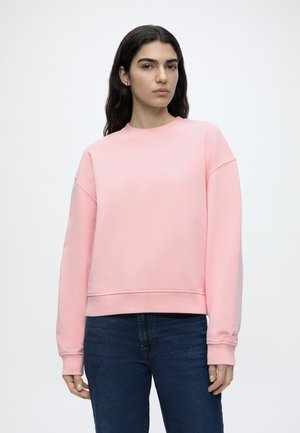 Kobieta o długich ciemnych włosach, ubrana w jasnoróżową bluzę i ciemnoniebieskie dżinsy, stojąca na tle jednolitego jasnego tła.