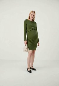 Oliwkowa dopasowana sukienka z długimi rękawami i z przodu z węzłem, zestawiona z małą kremową torebką i czarnymi płaskimi butami. Gładka tekstura.