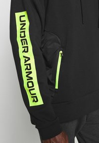 Felpa nera con "UNDER ARMOUR" in verde brillante sulla manica. Presenta una tasca laterale con una cerniera neon e un design elegante e testurizzato.
