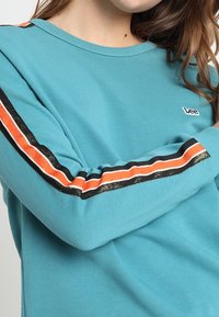 Maglietta a maniche lunghe turchese con dettagli a strisce nere, arancioni e bianche lungo le maniche. Presenta un piccolo logo "Lee" sul petto.