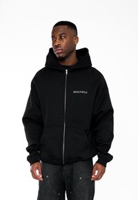 Felpa nera con zip realizzata in tessuto morbido, dotata di un ampio cappuccio e la parola "MULTIPLY" in grigio tenue sul petto. Design a vestibilità standard.