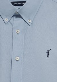 Ljust blå skjorta med krage i bomull, med vita knappar och en liten marinblå broderad figur på vänster bröst.