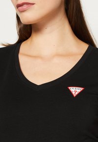 Femme portant un t-shirt noir à col en V avec un petit logo Guess rouge et blanc sur la poitrine gauche.