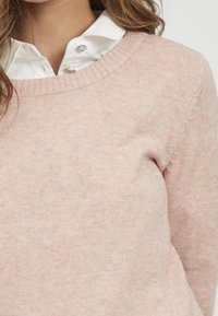 Ljusrosa stickad tröja med bred rund halsringning, mjuk textur och ribbade ärmslut, kombinerad med en vit skjorta med krage under.