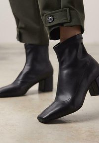 Botas de cuero negras hasta el tobillo con punteras puntiagudas, tacones de bloque y un diseño cosido. Las botas presentan una textura suave y una parte superior ajustada.