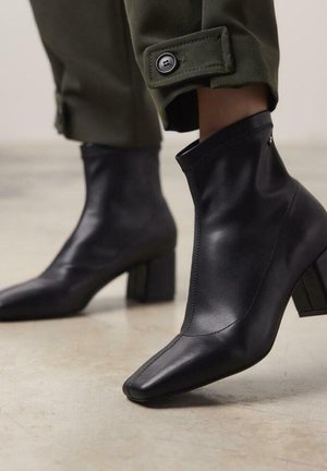 Botas de cuero negras hasta el tobillo con punteras puntiagudas, tacones de bloque y un diseño cosido. Las botas presentan una textura suave y una parte superior ajustada.