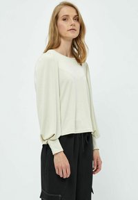 Maglietta a manica lunga color crema con scollatura rotonda, caratterizzata da maniche a sbuffo e una vestibilità rilassata, abbinata a pantaloni neri.