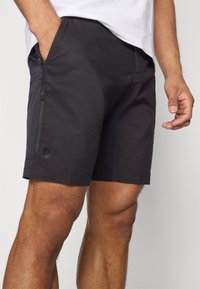 Schwarze Shorts aus leichtem Stoff, mit einer Reißverschlusstasche, einem Logo am linken Bein und einem einfachen, schlanken Design.
