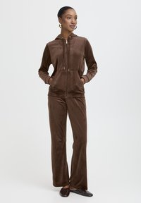 Ensemble de survêtement en velours marron comprenant un hoodie zippé avec poches, un pantalon assorti à coupe décontractée et une texture lisse. Légère brillance visible.