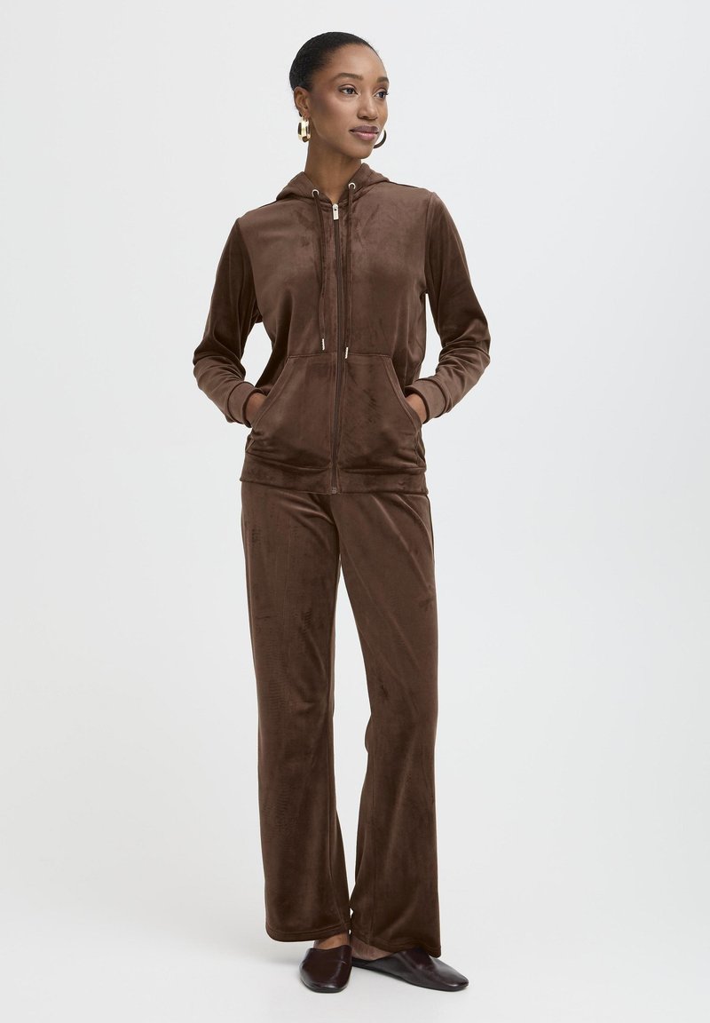 Ensemble de survêtement en velours marron comprenant un hoodie zippé avec poches, un pantalon assorti à coupe décontractée et une texture lisse. Légère brillance visible.